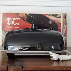 May include: A vintage electric hot server by Cornwall, with a black, domed lid and a rectangular base. The server is on a wooden surface, with the original box in the background. The box has the words "electric hot server" printed on it.