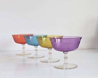 May include: A set of four stemmed coupe glasses featuring translucent bowls in orange, blue, yellow, and purple. Each glass has a wide, shallow bowl with a textured surface and a clear, circular base supported by a short, multi-tiered stem.