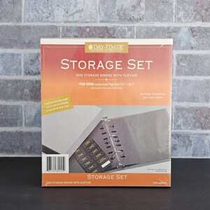 May include: A Day-Timer Storage Set, featuring a binder with a slipcase. The packaging is primarily orange and white, with the text "STORAGE SET" prominently displayed. Includes sheetlifter and year labels. The binder is designed for 14 cm x 22 cm pages.
