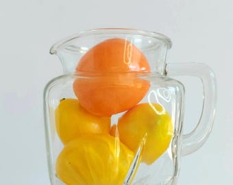 May include: A clear glass pitcher with a square base and a side handle, containing one large round orange fruit and three smaller yellow citrus fruits inside the transparent vessel against a plain white background.