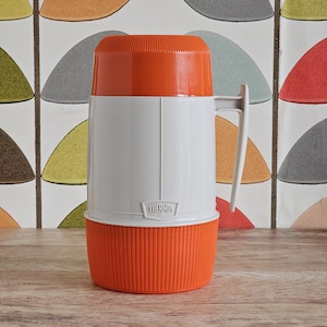 May include: A retro Thermos flask in white and orange. The flask has a white handle and the Thermos logo is visible. The top and bottom sections are orange, while the main body is white. The background features a retro pattern.