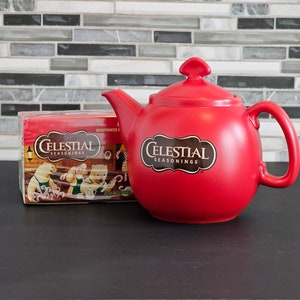 May include: A red ceramic teapot with a white "Celestial Seasonings" logo on the front. A box of Celestial Seasonings tea is in the background.