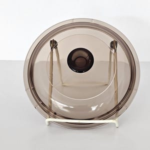 May include: A round, transparent, smoke-coloured glass lid featuring a central circular knob and a metal wire handle mechanism attached to the sides. The lid is positioned upright, showcasing its smooth, curved surface and dark, translucent tint.