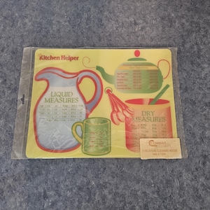 Peut inclure: Guide de mesure Kitchen Helper jaune et rouge avec un pichet, une tasse et un bol. Le guide comprend des mesures liquides et sèches. Le texte sur le guide indique "Kitchen Helper", "Liquid Measures" et "Dry Measures".