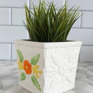 May include: A square white ceramic planter with a basket-weave texture and a rope-style rim. One side features a painted orange flower with yellow accents and green leaves, while the other side displays a matching embossed floral design. Contains grass.