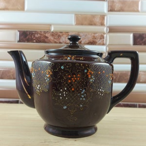 May include: A brown ceramic teapot with a gold trim and floral design in shades of orange, blue, and white.