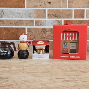 May include: A selection of Tim Hortons Christmas ornaments. Features a black coffee pot ornament with the text "Tim Hortons 12:25", a snowman ornament, and a red box with a store front ornament.
