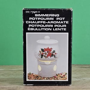 May include: A white ceramic potpourri pot with a floral design, presented in its original black box. The box has text in English and French, including "Simmering Potpourri Pot". The pot has a lid and a base with openings for the fragrance.