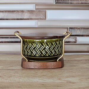 Peut inclure: Un plat de service vintage avec un bol en céramique vert foncé orné d'un motif tissé. Il possède un couvercle et une base en bois, avec des poignées en métal doré. Le plat est conçu pour servir des aliments.