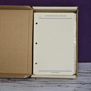 May include: A cream-coloured, three-hole punched correspondence and notes refill page for a binder, featuring horizontal lines, a decorative border, and the text "Correspondence/Notes" and "The Priority Manager" inside a cardboard box.