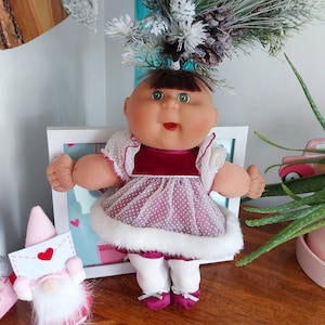 May include: A Cabbage Patch Kids doll wearing a white and pink dress with a white fur trim. The doll has a red velvet bodice and green eyes. The doll is wearing white tights and pink shoes. The doll is wearing a festive holiday outfit.