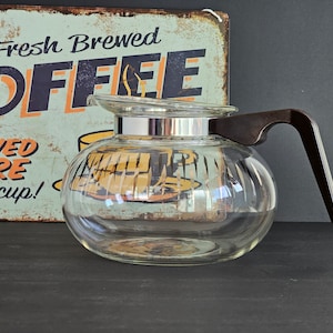 May include: A clear glass coffee pot with a brown plastic handle. The pot has a silver band around the top and a textured glass body. The pot is sitting on a black surface in front of a vintage metal sign that reads "Fresh Brewed Coffee 25¢ Served Here Have a cup!"