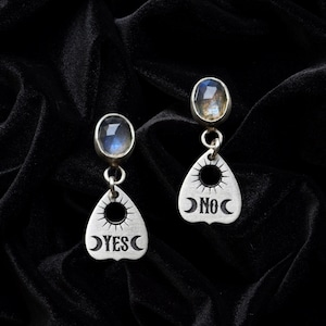 May include: A pair of silver-toned earrings. Each earring features a teardrop-shaped charm with the words "YES" and "NO" and a crescent moon design. The earrings have a small, oval-shaped gemstone at the top.