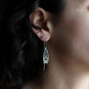 Peut inclure: Boucles d'oreilles chauve-souris en argent avec détails en émail noir. Les boucles d'oreilles sont suspendues à une chaîne délicate, présentant un motif de chauve-souris stylisé. Les ailes de la chauve-souris sont noires, et le corps est argenté.