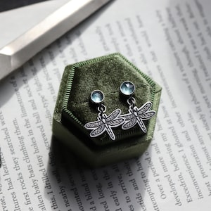 Könnte beinhalten: Silberne Ohrringe mit Libellen-Motiv und hellblauen Edelstein-Nieten, präsentiert auf einer grünen Samt-Schmuckschatulle. Die Ohrringe haben detaillierte Flügeldesigns, vor einem Hintergrund mit gedrucktem Text.
