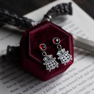 Könnte beinhalten: Silberne Ohrringe mit einem Marienkäfer-Design. Jeder Ohrring hat einen kleinen, runden, roten Edelstein und einen baumelnden Marienkäfer-Anhänger mit schwarzen Punkten. Präsentiert auf einem bordeauxroten Samt-Schmuckkästchen.