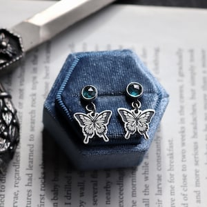 Könnte beinhalten: Silberne Schmetterlingsohrringe mit teal-farbenen Edelsteinen, präsentiert auf einer blauen Samtbox. Die Ohrringe haben ein detailliertes Schmetterlingsdesign. Ein zartes Accessoire.