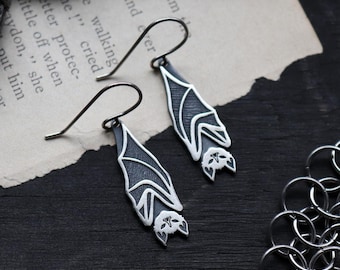 May include: A pair of silver bat earrings with a dark gray textured finish. The bat wings have white outlines, and the bat's face is detailed. The earrings hang from curved ear wires, showcasing a gothic or Halloween theme.
