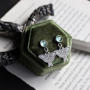 Könnte beinhalten: Silberfarbene Ohrringe mit einem detaillierten Motten-Design, jeweils mit einem kleinen, runden, hellgrünen Edelstein. Die Ohrringe werden in einer dunkelgrünen Samtbox präsentiert, mit einem Vintage-Buch im Hintergrund.