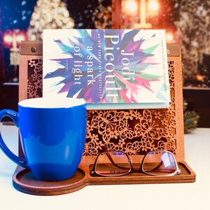 May include: A wooden book stand with a blue mug, a pair of glasses, and a book titled "a spark of light" by Jodi Picoult. The stand has a decorative laser-cut design with butterflies and flowers.