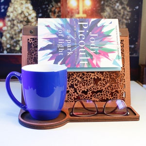 May include: A wooden book stand with a blue mug, a book titled "a spark of light" by Jodi Picoult, and a pair of glasses. The stand has a decorative butterfly design.