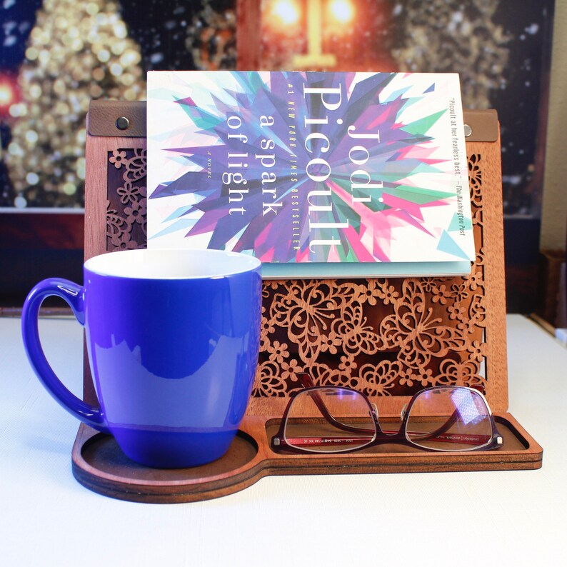 May include: A wooden book stand with a blue mug, a book titled "a spark of light" by Jodi Picoult, and a pair of glasses. The stand has a decorative butterfly design.