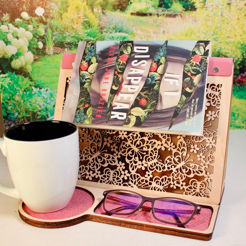May include: A wooden book stand with a pink base and a cutout design of butterflies and flowers. A book titled "If I Disappear" by Lia Jane Brazier is resting in the stand. A white mug and a pair of glasses are on the stand.