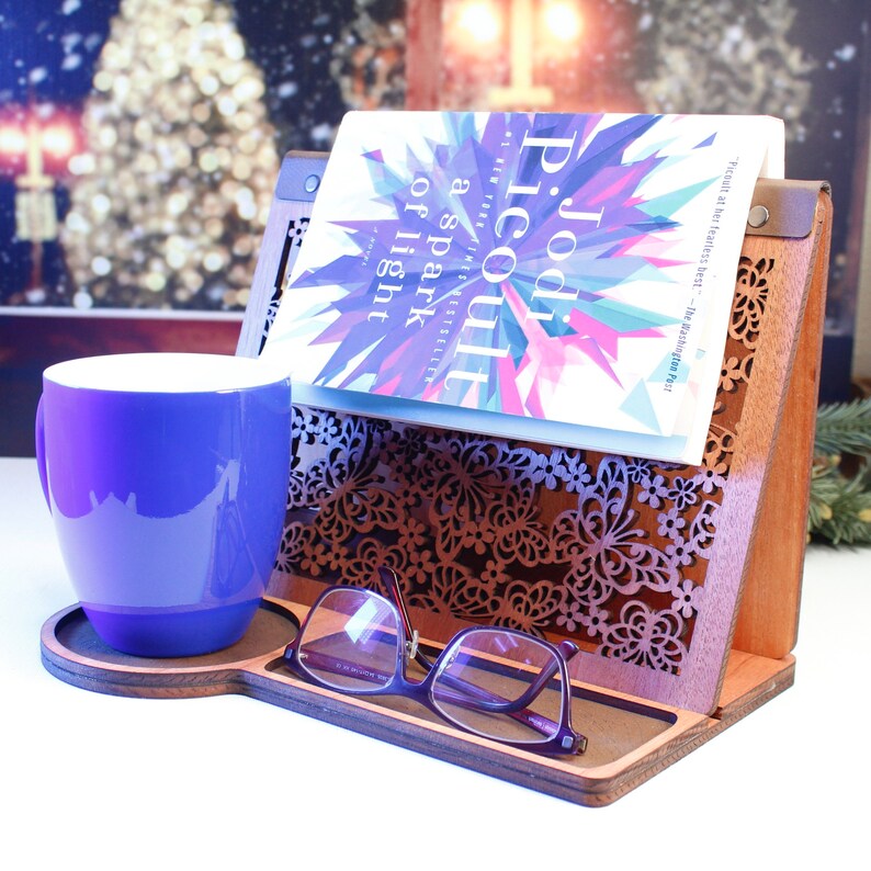 May include: A wooden book stand with a laser-cut butterfly design. A book titled "A Spark of Light" by Jodi Picoult is resting on the stand. A purple mug and glasses are on the stand.