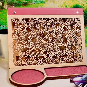 May include: A wooden organizer with a pink top and a cutout design of butterflies and flowers. The organizer has two compartments, one with a pink circle and the other with a pink rectangle.