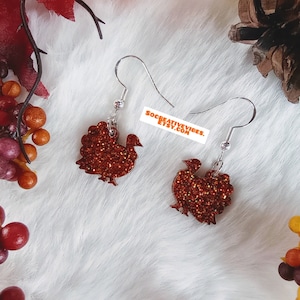 May include: A pair of dangle earrings featuring a red glitter turkey design. The earrings are made with silver hooks.