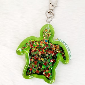 May include: A green turtle-shaped keychain with a silver clasp. The turtle is filled with colorful glitter in shades of red, green, and gold. The keychain is made of resin and has a glossy finish. The background is a white, fluffy surface.