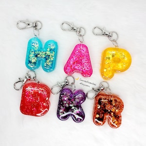 May include: A collection of colorful resin letter keychains. The letters are filled with glitter and small decorative elements. Each keychain has a silver clasp. The letters are in various colors, including blue, pink, yellow, red, purple, and orange.