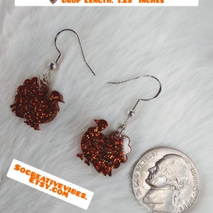 May include: A pair of dangle earrings featuring a glittery orange turkey silhouette. The earrings are 5/8 inch wide and tall, with a drop length of 1.25 inches. "TURKEY WIDTH AND HEIGHT 5/8" OF AN INCH DROP LENGTH: 1.25" INCHES" is written in orange text at the top of the image. "SOCREATIVEVIBES. ETSY.COM" is written in orange text at the bottom of the image.
