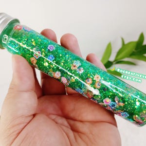May include: A clear glass tube filled with green glitter and small, colorful flower-shaped confetti. The tube has a silver cap and is held in a hand. The text "SOCREATIVEVIBES.ETSY.COM" is visible.