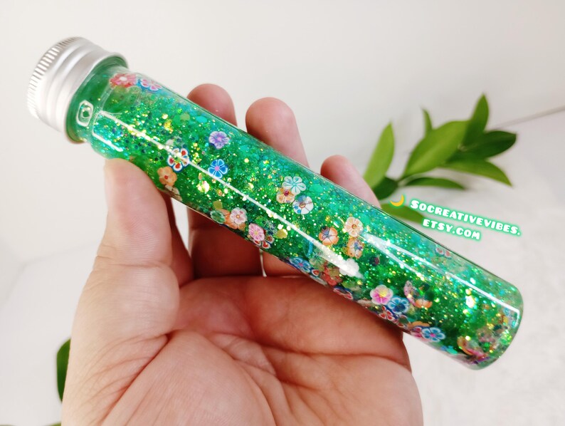May include: A clear glass tube filled with green glitter and small, colorful flower-shaped confetti. The tube has a silver cap and is held in a hand. The text "SOCREATIVEVIBES.ETSY.COM" is visible.