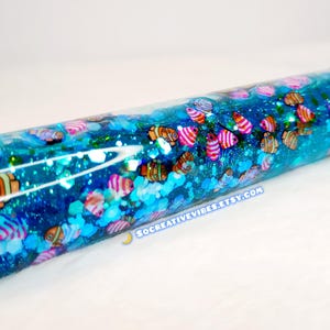 May include: A blue glitter tumbler with a black lid, filled with blue liquid and colorful fish-shaped decorations. The tumbler has a clear, glossy finish and the text "SOCREATIVEVIBES.ETSY.COM" is visible.