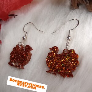 May include: A pair of dangle earrings featuring a turkey silhouette made of orange glitter resin. The earrings have silver hooks.