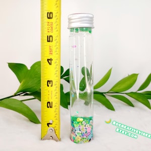 May include: A clear glass tube with a silver cap, filled with colorful confetti and glitter. The tube is approximately 4 inches tall. Green leaves and a yellow measuring tape are in the background. The Etsy shop name is visible.