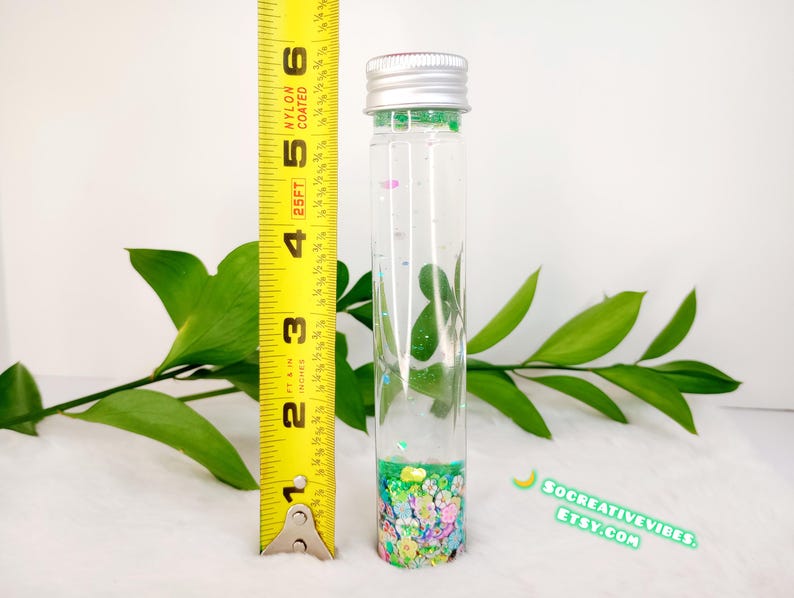 May include: A clear glass tube with a silver cap, filled with colorful confetti and glitter. The tube is approximately 4 inches tall. Green leaves and a yellow measuring tape are in the background. The Etsy shop name is visible.