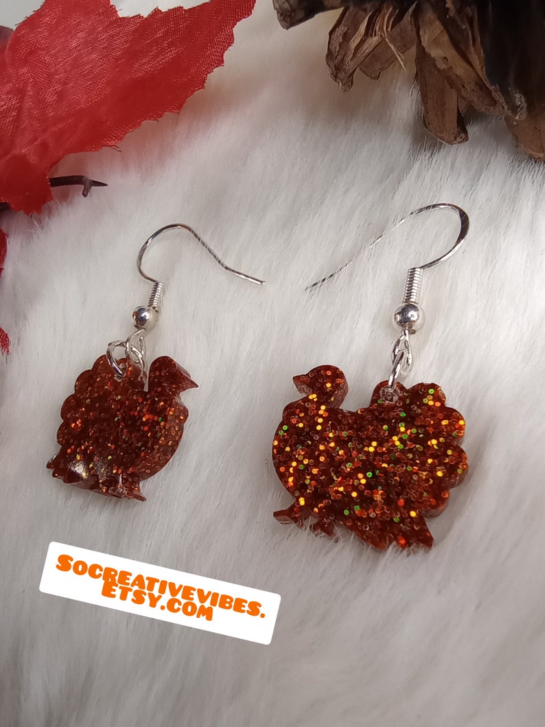 May include: A pair of dangle earrings featuring a turkey silhouette made of orange glitter resin. The earrings have silver hooks.
