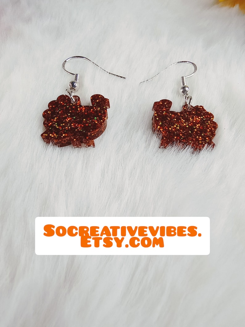 May include: A pair of brown glitter turkey earrings with silver hooks. The earrings are on a white fuzzy background.