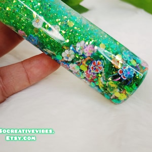 May include: A clear, cylindrical bottle filled with green glitter and colorful floral shapes. The bottle is held against a white surface. The text "SOCREATIVEVIBES. ETSY.COM" is visible at the bottom.