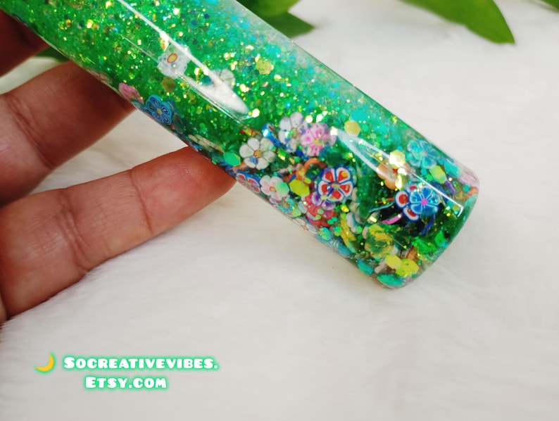 May include: A clear, cylindrical bottle filled with green glitter and colorful floral shapes. The bottle is held against a white surface. The text "SOCREATIVEVIBES. ETSY.COM" is visible at the bottom.