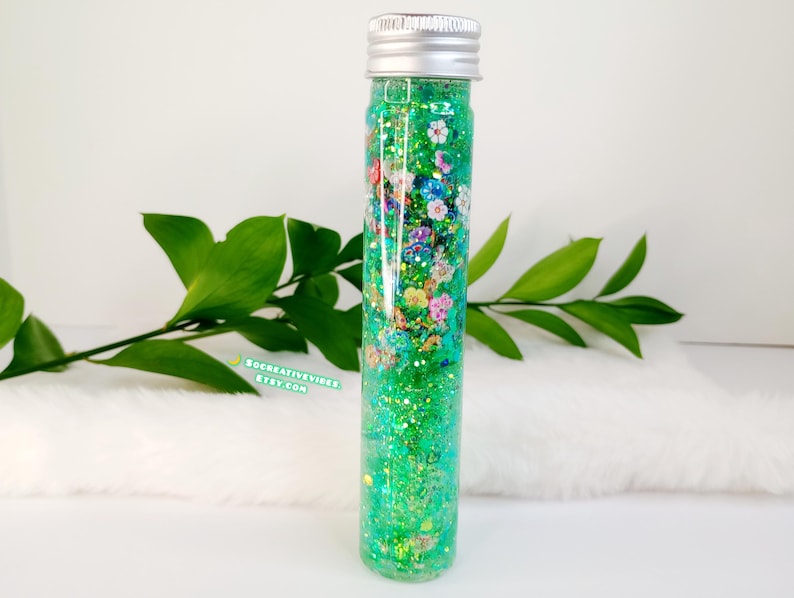 May include: A clear glass tube filled with green glitter, small flowers, and silver accents. The tube has a silver cap and is surrounded by green leaves. The text "SOCREATIVEVIBES.ETSY.COM" is visible.