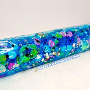 May include: A cylindrical, clear water bottle filled with blue liquid, glitter, and colorful fish-shaped confetti. The bottle has a dark cap and the text "SOCREATIVEVIBES.ETSY.COM" is visible.