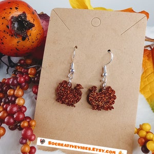 May include: A pair of dangle earrings featuring a glittery red and orange turkey design. The earrings are on a brown card with the text "socreativevibes.etsy.com".