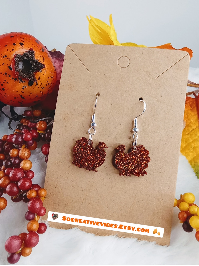 May include: A pair of dangle earrings featuring a glittery red and orange turkey design. The earrings are on a brown card with the text "socreativevibes.etsy.com".