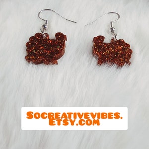 May include: A pair of brown glitter turkey earrings with silver hooks. The earrings are on a white fuzzy background.