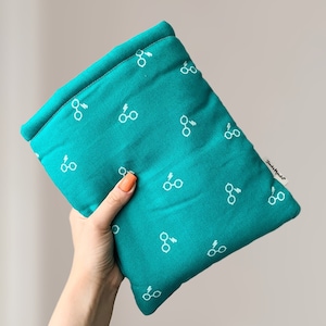May include: Teal rectangular cushion featuring a white glasses and lightning bolt pattern. The cushion is folded over and held in a hand. The brand name "Jude & Mele" is printed on a small white tag.
