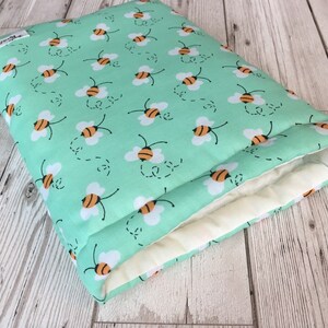 May include: A rectangular, mint green fabric item with a repeating pattern of cartoon bees. The bees are yellow and black with white wings. The interior is a cream color. The item appears to be a pillow or a case.